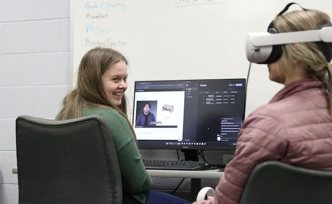 Two students interact as one sits in front of a computer screen and the other wears a VR headset.