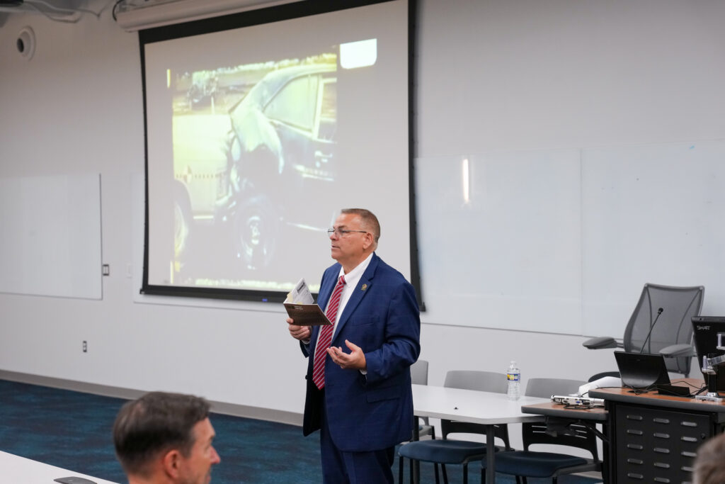 A man in a suit standing in a room, holding a book and giving a lecture to an audience.