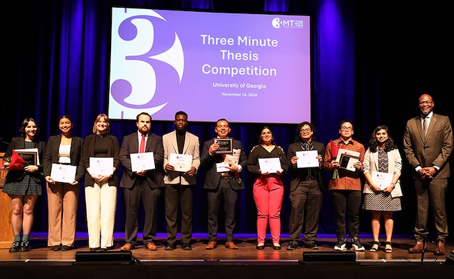 group of students holding awards in front of Three Minute Thesis sign on stage