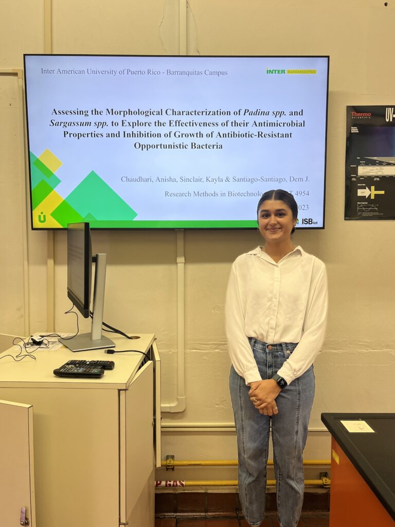 Anisha stands in front of academic presentation on digital television screen
