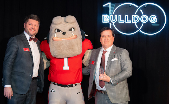 Justin Greer and Frankin Pittman stand in front of the Bulldog 100 neon sign with their award and Hairy Dawg