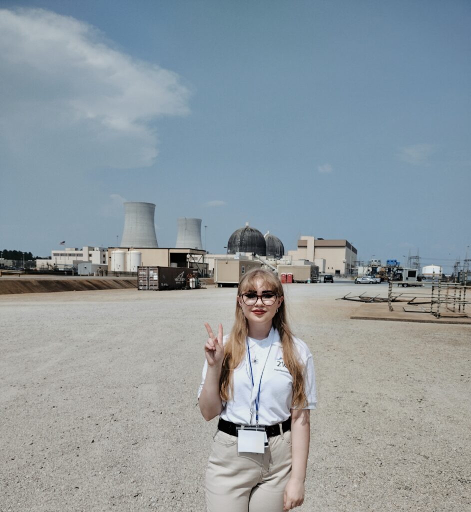 Katie stands making a peace sign in front of a power plant